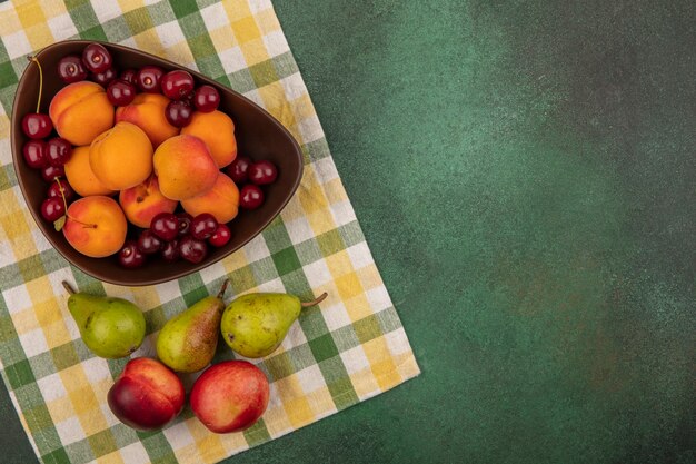 ボウルにアプリコットとチェリーとして果物の上面図とコピースペースと緑の背景に格子縞の布に梨と桃のパターン