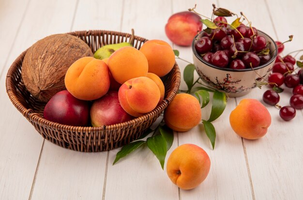 Foto gratuita vista dall'alto di frutta come noce di cocco albicocca pesca pera nel cestino e ciotola di ciliegie con foglie su sfondo di legno