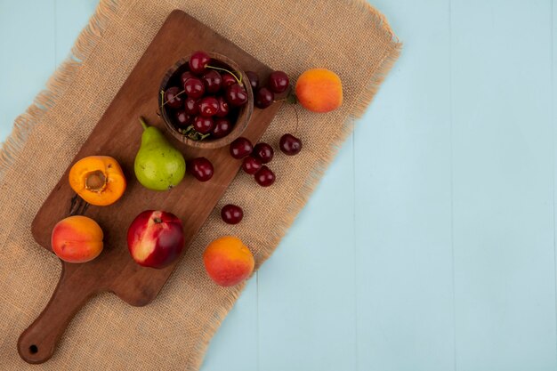 Top view of fruits as apricots peach pear and bowl of cherry on cutting board and on sackcloth on blue background with copy space