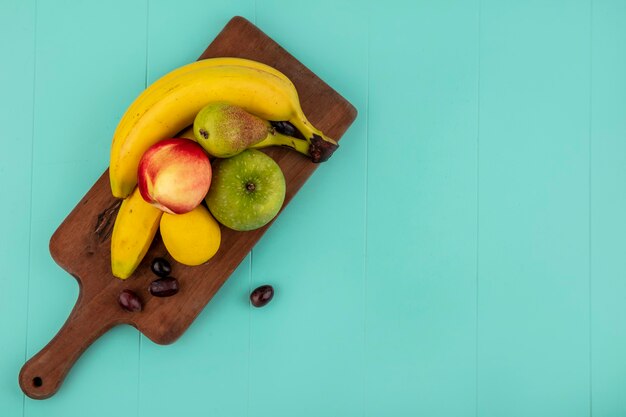 Top view of fruits as banana apple lemon peach grape berries on cutting board on blue background with copy space