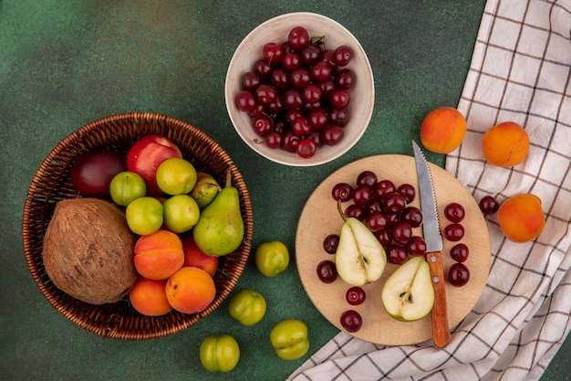 Top view of fruits as cherries pear coconut plum apricot peach with knife in basket and on cutting board on plaid cloth on green background