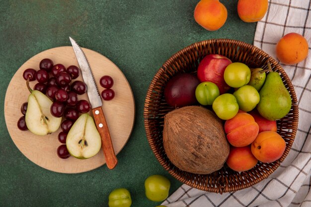 Top view of fruits as cherries pear coconut plum apricot peach with knife in basket and on cutting board on plaid cloth on green background