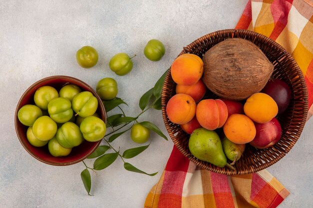 Top view of fruits as coconut apricot peach pear in basket on plaid cloth with bowl of plums on white background