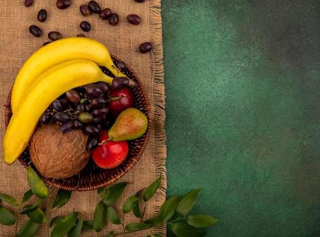 Foto gratuita vista dall'alto di frutta come cocco banana uva pera pesca nel cesto con foglie su tela di sacco su sfondo verde con spazio di copia