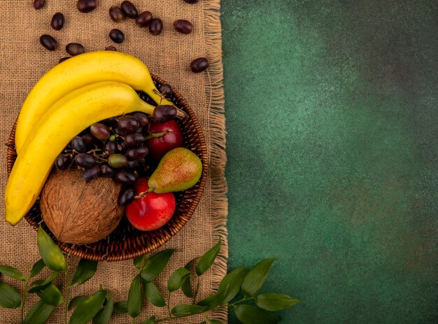 Вид сверху на фрукты как кокос, банан, виноград, груша, персик в корзине с листьями на мешковине на зеленом фоне с копией пространства