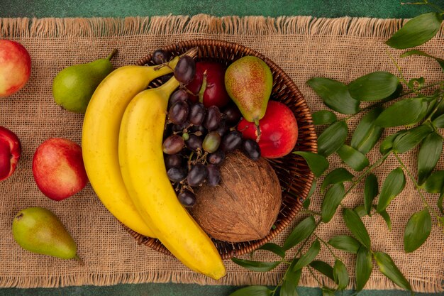 Top view of fruits as coconut banana grape pear peach in basket with leaves on sackcloth on green background