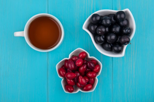 Vista dall'alto di frutti come bacche di corniolo e prugnole in ciotole con tazza di tè su sfondo blu