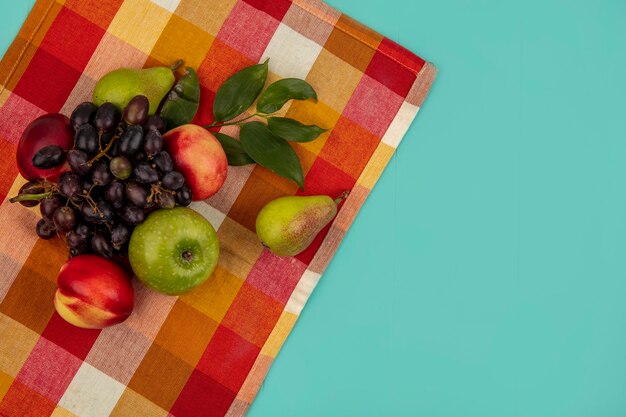 Top view of fruits as grape peach apple pear with leaves on plaid cloth and blue background with copy space