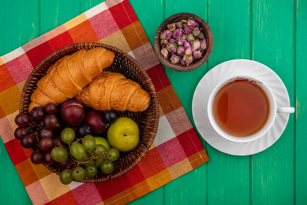 Top view of fruits as grape pluots sloe berries with croissants in basket on plaid cloth and bowl of flowers with cup of tea on green background