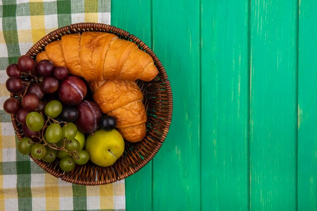 格子縞の布とコピースペースと緑の背景の上のバスケットにクロワッサンとブドウのプルオットスローベリーとしての果物の上面図