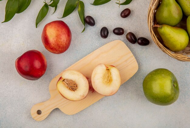 Top view of fruits as half cut peach on cutting board and bowl of pear with grape berries apple with leaves on white background