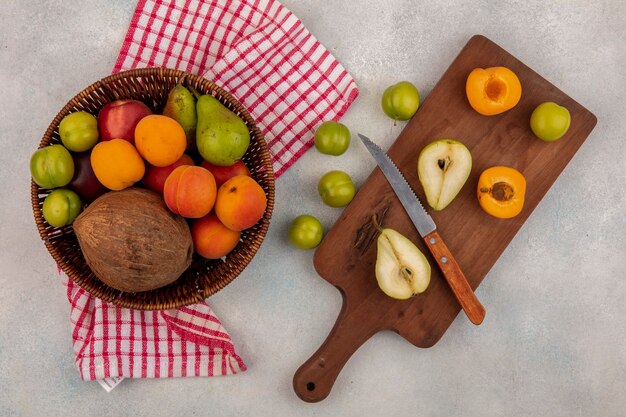 Top view of fruits as half cut pear and apricot with knife on cutting board and basket of coconut peach plum pear on plaid cloth and plums on white background