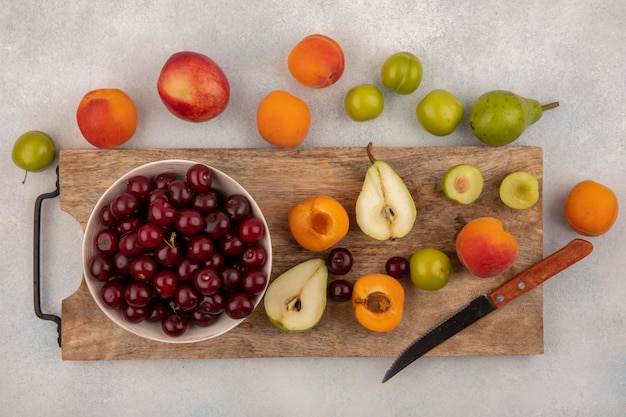 Top view of fruits as half cut pear plum apricot with knife and bowl of cherry on cutting board and pattern of pear plum apricot on white background