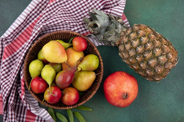緑の表面にザクロとパイナップルの格子縞の布の上のバスケットに桃リンゴプラムとして果物のトップビュー