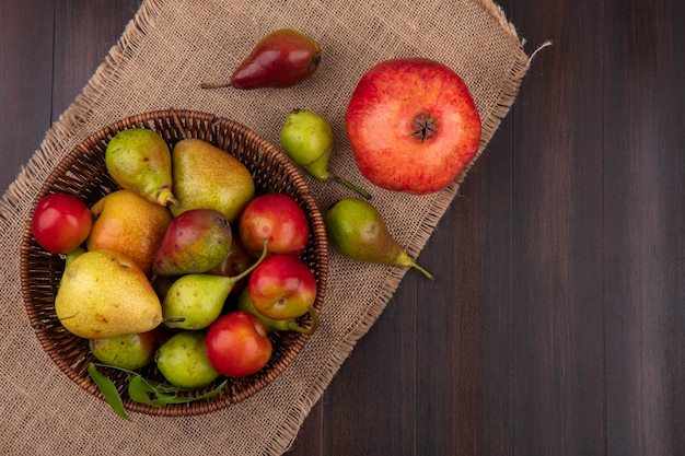 Top view of fruits as peach apple plum in basket with pomegranate on sackcloth on wooden surface