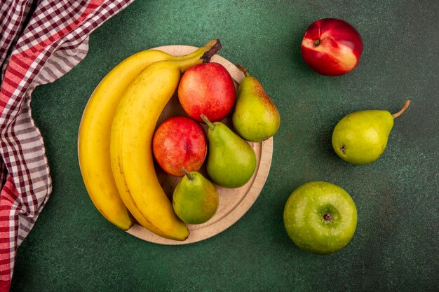 Top view of fruits as peach pear banana on cutting board with apple and plaid cloth on green background