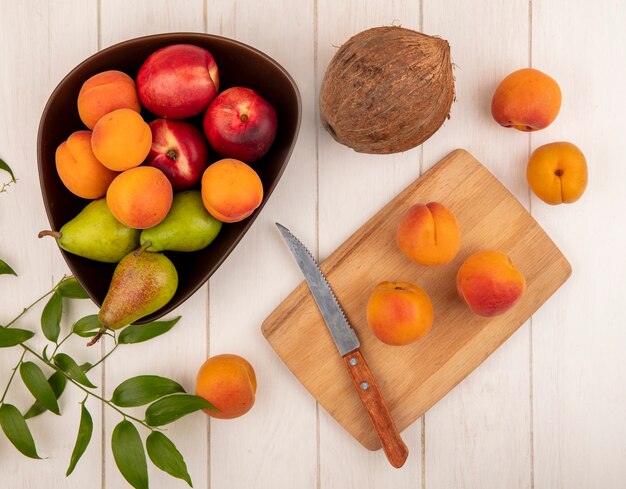 Top view of fruits as peach pear in bowl and peaches with knife on cutting board with coconut and leaves on wooden background