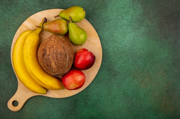 Top view of fruits as peach pear coconut banana on cutting board on green background with copy space