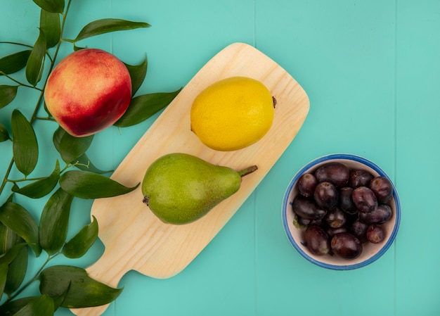 Вид сверху на фрукты в виде груши и лимона на разделочной доске и миску с виноградными ягодами и персиком с листьями на синем фоне