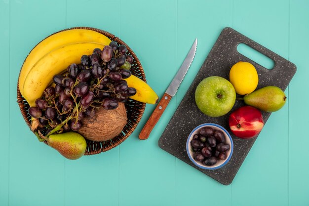 Вид сверху на фрукты, такие как груша, персик, яблоко и виноградные ягоды, лимон на разделочной доске и корзина из бананового кокосового винограда с ножом на синем фоне