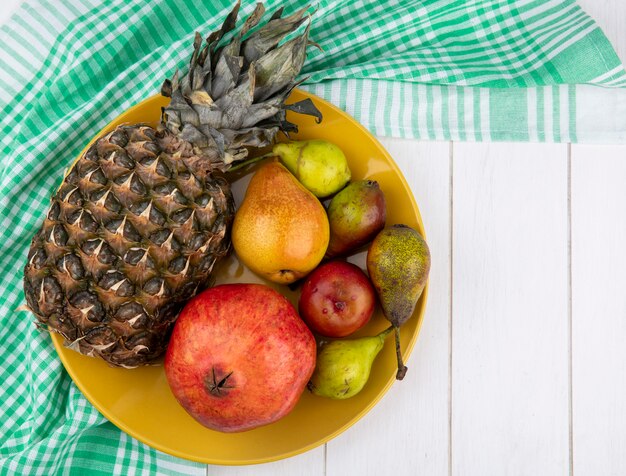 Top view of fruits as pineapple, pears, pomegranate and peach in plate on plaid cloth on wooden surface with copy space