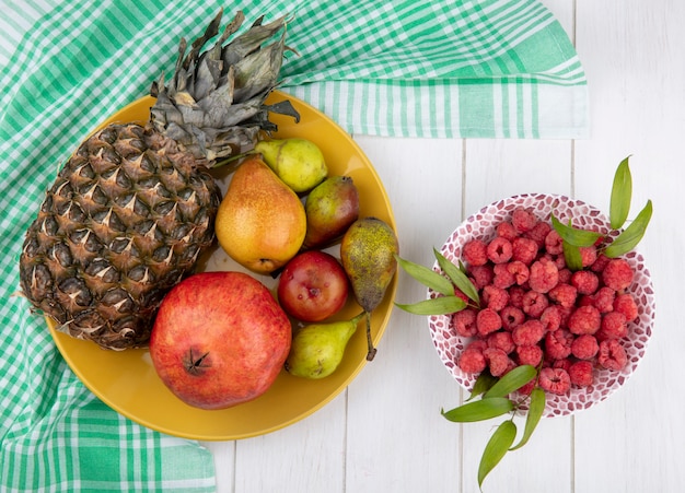 Top view of fruits as pineapple pomegranate peach in plate on plaid cloth with raspberries in bowl on wooden surface