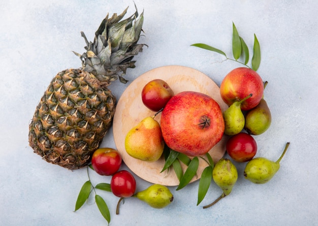 Вид сверху на фрукты, сливы, персик и гранат на разделочной доске и с яблоком и ананасом на белой поверхности