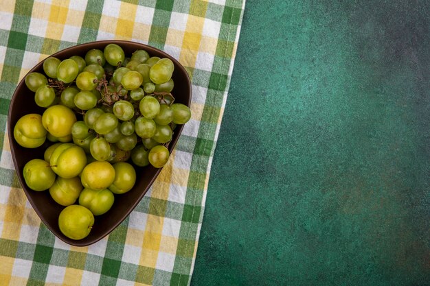 Top view of fruits as plums and grape in bowl on plaid cloth and green background with copy space