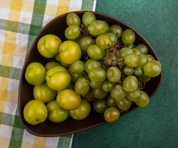 Top view of fruits as plums and grape in bowl on plaid cloth and green background