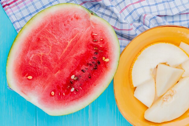 Top view of fruits as watermelon half and plate of melon slices on plaid cloth and blue background