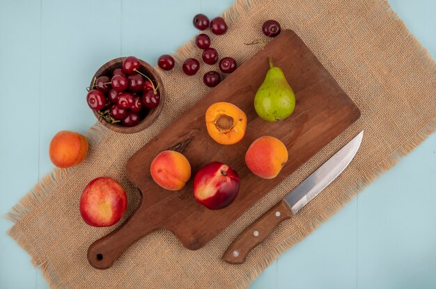 Top view of fruits as whole and half apricots and peach pear on cutting board and cherries in bowl and peach apricot with knife on sackcloth and blue background