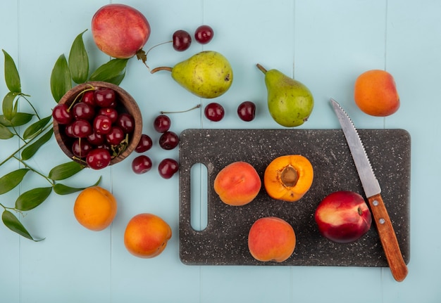 Вид сверху на фрукты в целом и половину абрикосов и персика с ножом на разделочной доске и вишни в миске и узор из груши, персика, абрикоса, вишни с листьями на синем фоне