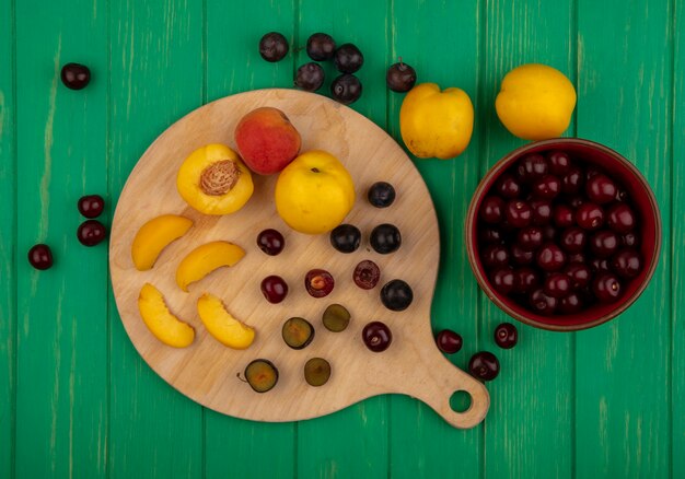 Top view of fruits as whole half sliced apricots and sloe berries with knife on cutting board and bowl of cherries on green background