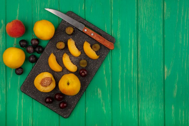 Top view of fruits as whole half sliced apricots and sloe berries with knife on cutting board and on green background with copy space