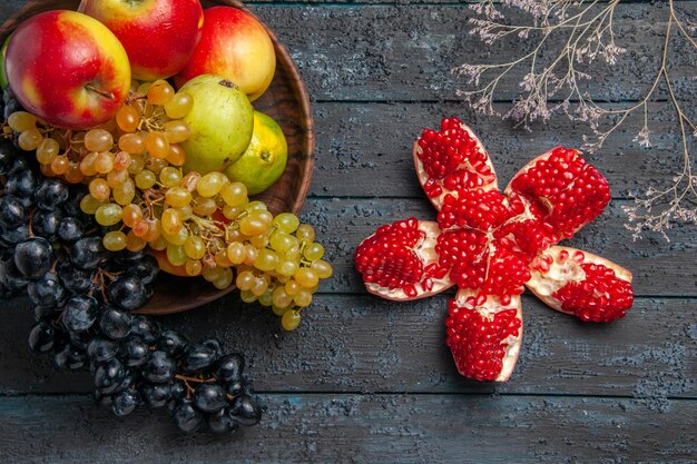 Вид сверху фрукты в миске, миска с белым и черным виноградом, лаймами, яблоками, грушами, рядом с гранатом и ветками на серой поверхности
