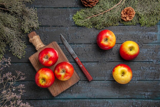 Вид сверху фрукты и нож три желто-красноватых яблока на деревянной разделочной доске рядом с ножом и три яблока под ветвями деревьев с шишками на столе