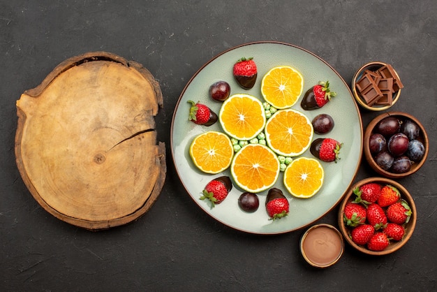 Top view fruits on table strawberries chocolate and berries in wooden bowls next to the plate of chopped orange candies and chocolate-covered strawberries next to wooden kitchen board