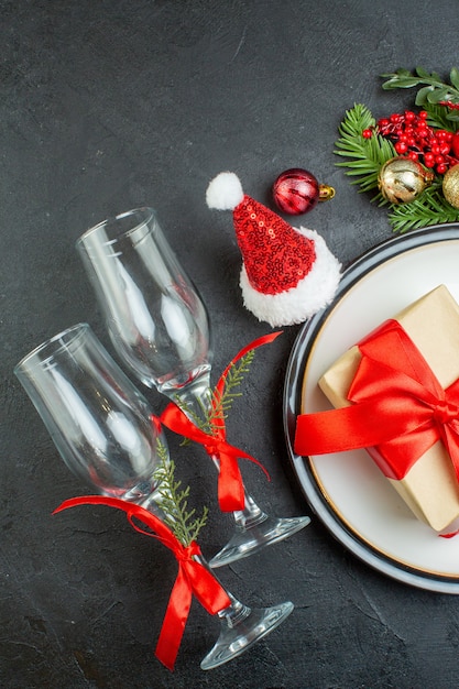 Top view of gift box on dinner plate christmas tree fir branches conifer cone santa claus hat fallen glass goblets on dark background