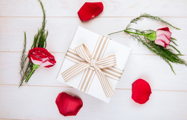 Top view of a gift box tied with bow and red color roses with scattered petals and asparagus on white wooden background