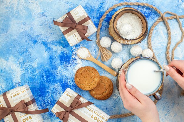 Top view gift boxes coconut balls rope cookies cup of milk in female hand on blue white background