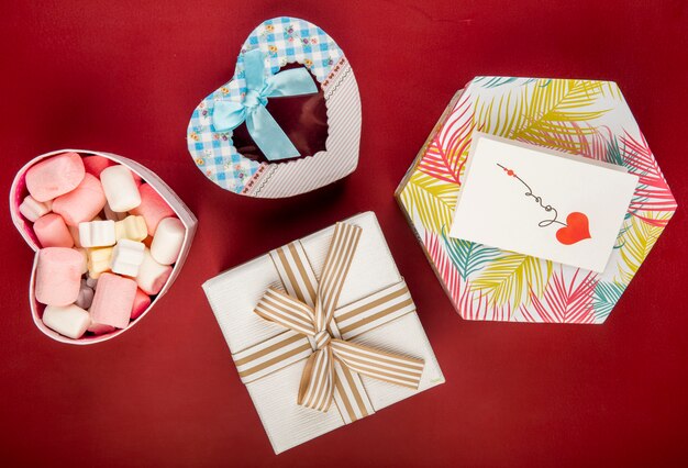 Top view of gift boxes of different shapes and colors and marshmallow in a heart shaped box on red table