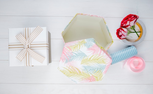 Top view of gift boxes and red color roses with rolls of adhesive tape and pink ribbon on white wooden background