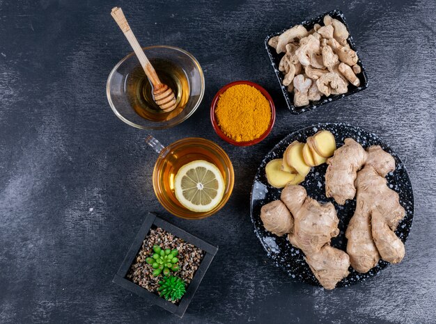 Top view ginger in bowls and plate with honey, a cup of tea with lemon, ginger slices and powder on dark textured background. horizontal