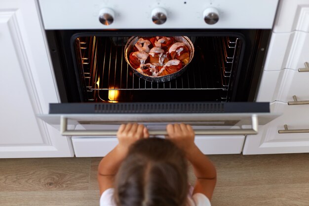 Top view of girl child opening oven looking at tasty croissant or other baking n the oven, cooking process on kitchen, small dark haired female kid wants delicious sweets.