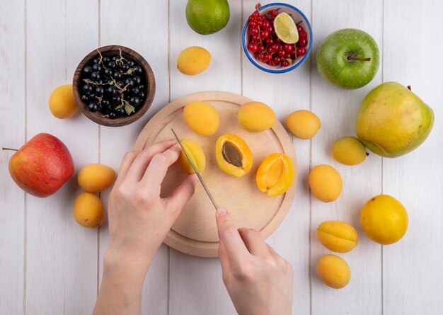 Top view of a girl cuts apricots on a stand with apples and black and red currants on a white surface