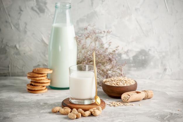 Top view of glass bottle and cup filled with milk on wooden tray and dry fruits stacked cookies spoon oats in brown pot on the left side on white table on ice background