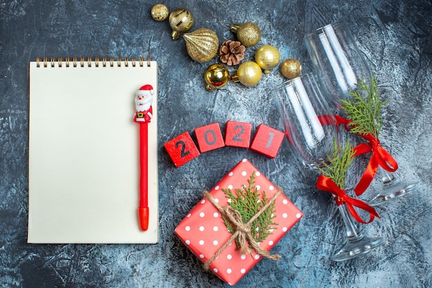 Top view of glass goblets with red ribbon and numbers decoration accessories next to gift boxes notebook with pen on dark table