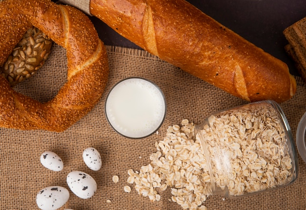 Vista dall'alto di bicchiere di latte e vaso sparso con fiocchi d'avena con pane su tela di sacco su sfondo nero