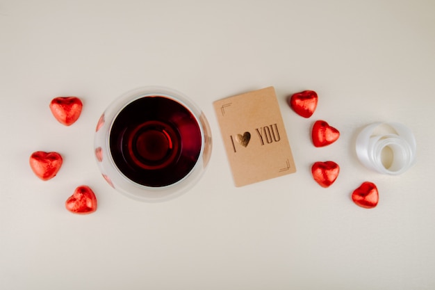 Top view of a glass of wine with heart shaped chocolate candies wrapped in red foil and a small postcard on white table