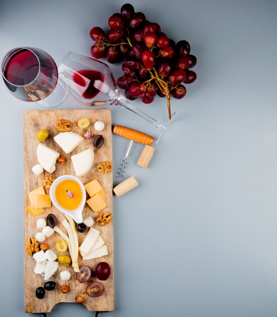 Top view of glasses of red wine with cheese grape olive nuts butter on cutting board and corkscrew on white with copy space